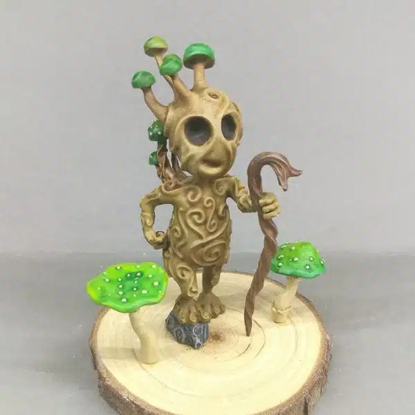 Kodama gardien de champignons sur son socle en bois. Il s'agit d'une figurine en résine représentant un esprit des bois marchant vaillamment, un bâton à la main. Autour de lui se trouvent 2 champignons verts.