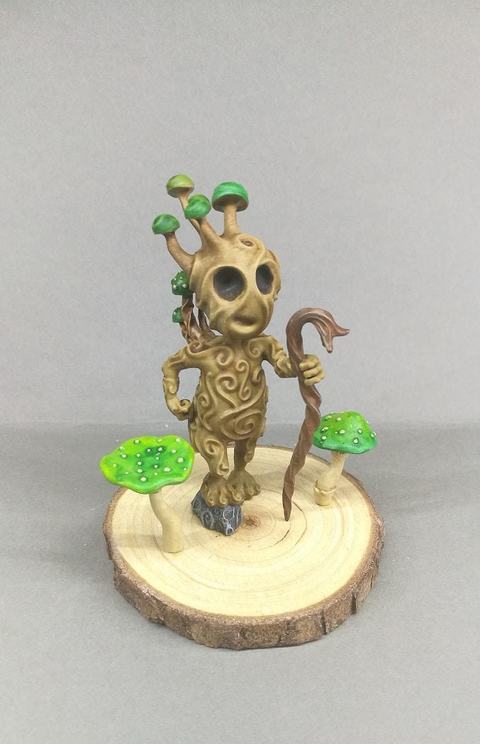 Kodama marcheur sur son socle en bois. Il s'agit d'une figurine en résine représentant un esprit des bois marchant vaillamment, un bâton à la main. Autour de lui se trouvent 2 champignons verts.