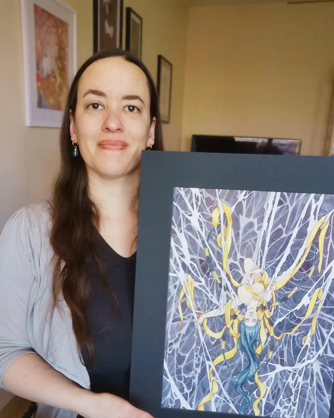 Un portrait de l'artiste Aemarielle tenant dans ses mains une de ses aquarelles originales issues de la collection Esprits japonais : Jorogumo. L'aquarelle représente une femme-araignée à l'envers dans une toile complexe. Elle est drapée de rubans de soie jaune.