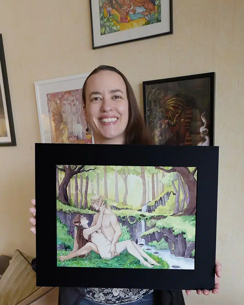 Un portrait d'Aemarielle qui tient dans ses mains une aquarelle personnalisée dans un passepartout noir. L'aquarelle représente 2 elfes enlacés, allongés dans l'herbe. Derrière eux, de la forêt, une cascade et une rivière.