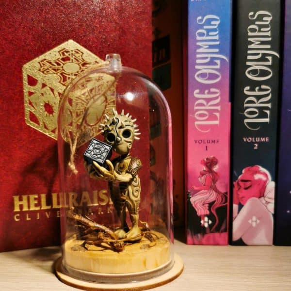 Un kodama Tête d'épines en résine, sous une cloche transparente, posé devant le célèbre roman Hellraiser. La figurine tient une mystérieuse boîte cubique dans les mains.