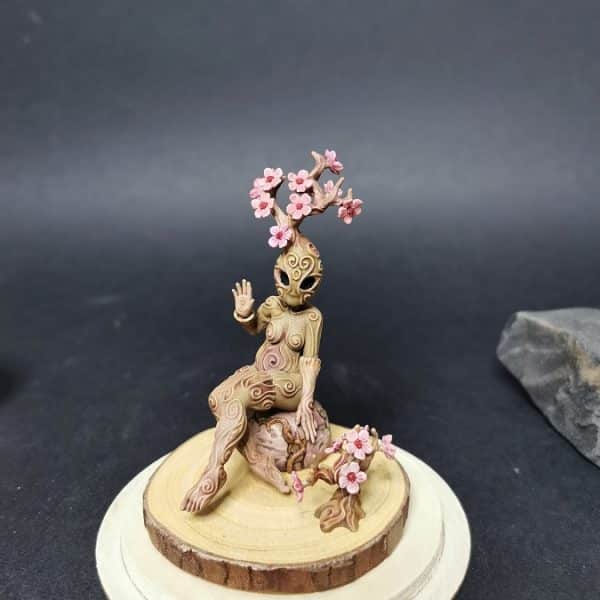 Une figurine en résine représentant une créature sylvestre coiffée de fleurs de cerisiers, assise sur un socle en bois.