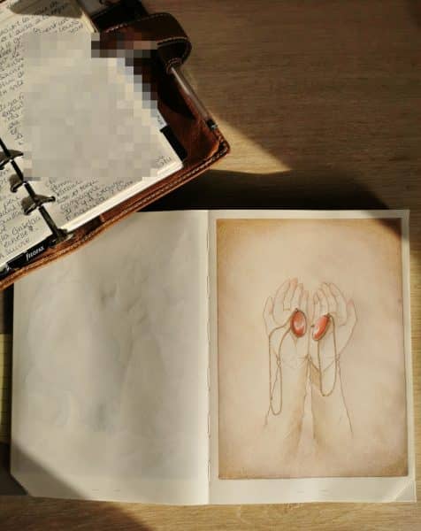 Le carnet à dessin ouvert sur deux mains tenant une amulette. À côté, le filofax sur lequel j'écris mes idées et premiers jets.