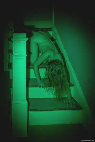Dans un escalier, une femme nue rampe en descendant les marches, les cheveux masquant son visage.