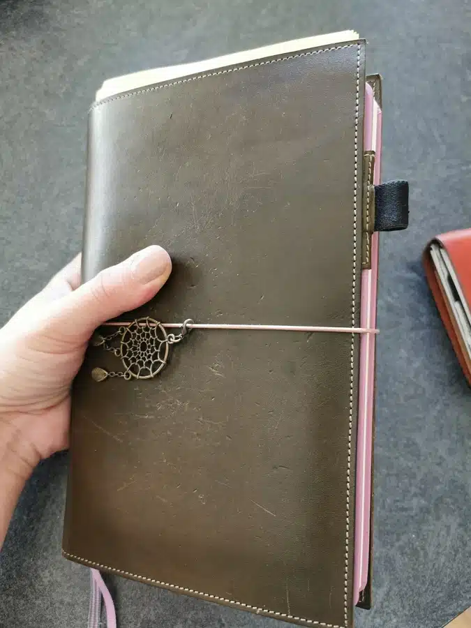 Couverture en cuir olive avec un carnet Leuchtturm A5 rose à l'intérieur. Un charme en laiton décore l'avant du carnet.